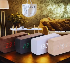 Pode incluir: Relógios digitais de madeira em várias cores, exibindo a hora com luzes LED. Os relógios têm formato retangular e são feitos de madeira em tons de marrom, preto, branco e madeira clara. A hora é exibida em vermelho, verde e branco.