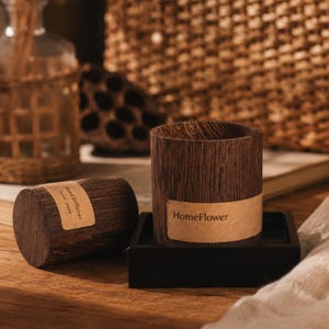 Puede incluir: Un recipiente cilíndrico de madera marrón oscuro con una etiqueta beige que dice HomeFlower se asienta sobre una base cuadrada negra. Junto a él hay un cilindro de madera a juego con una etiqueta beige que dice Reed Diffuser Since 21099.