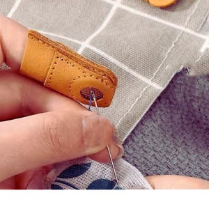 May include: Close-up of a leather finger protector for sewing. The tan leather thimble has a reinforced tip with a small hole for the needle. The thimble is on a finger, with a needle and thread piercing fabric.