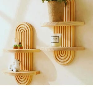Puede incluir: Dos estanterías de pared de madera con acabado natural. Cada estantería tiene un diseño curvo y arqueado con lamas verticales y una base semicircular. Una estantería contiene una planta en maceta, la otra una vela y objetos decorativos.