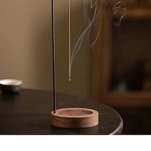 May include: Incense holder featuring a black metal stand and a wooden base. The incense stick is lit, with smoke rising. The holder is on a dark surface. The design suggests a focus on aromatherapy and relaxation, suitable for home use.