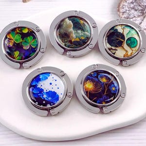 Könnte beinhalten: Fünf silberne Handtaschenaufhänger mit farbenfrohen, dekorativen Oberteilen. Die Designs umfassen ein vierblättriges Kleeblatt, einen bewölkten Himmel, einen Totenkopf, blaue Blumen und einen floralen Ausbruch. Diese zusammenklappbaren Handtaschenhaken sind zum Aufhängen von Taschen an Tischen bestimmt.