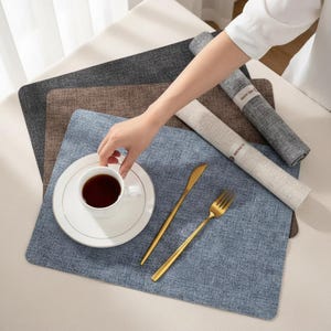 May include: A table setting with a white teacup and saucer, gold cutlery, and a blue placemat. Rolled-up grey and beige linen napkins are visible. Other placemats in grey and brown are also present.