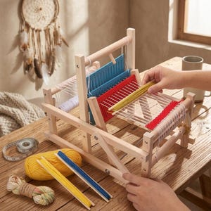 May include: A wooden weaving loom with red and blue guides, white and red woven threads, and yellow yarn. The loom is on a wooden table with a measuring tape and a dream catcher in the background. A person's hands are working with the loom.