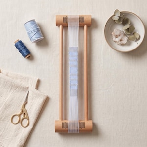 May include: A wooden weaving loom with white warp threads and a partially woven light blue fabric strip. Included are two spools of blue yarn, a pair of gold-toned scissors, and a ceramic bowl containing a crystal cluster and eucalyptus leaves.