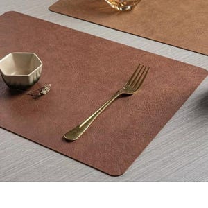 Peut inclure: Nappe rectangulaire marron avec une surface texturée. Une fourchette dorée repose sur le dessous de plat. Un petit bol hexagonal et une brindille de fleurs séchées sont également présents. Un autre dessous de plat et un verre sont en arrière-plan.
