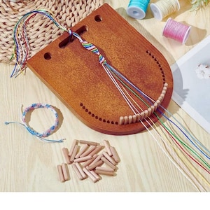 Könnte beinhalten: Ein hölzerner Armbandwebrahmen mit gebogenem Design, mit einem teilweise fertigen Armband und bunten Fäden. Zusätzliche Garnrollen in Blau, Rosa und Creme sind sichtbar, zusammen mit kleinen Holzstiften und einem fertigen Armband.