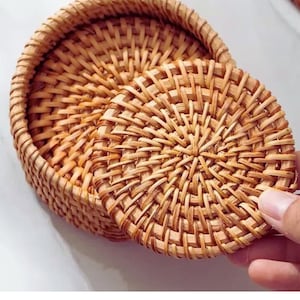 May include: A set of woven rattan coasters and a matching holder. The coasters are round with a tightly woven pattern, and the holder is a small basket. The natural tan color of the rattan gives a rustic, handmade feel.