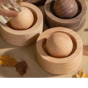 May include: Wooden aromatherapy diffusers in a light wood finish. Each diffuser has a round wooden base with a spherical wooden insert. A hand is shown holding a perfume bottle over one of the diffusers.