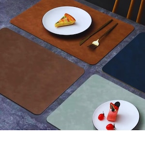 Puede incluir: Una mesa con manteles individuales rectangulares marrones, azules y verdes. Platos blancos con bordes negros muestran alimentos como quiche y pastel. Cubiertos dorados se colocan sobre el mantel individual marrón. La escena se desarrolla sobre una superficie oscura texturizada.