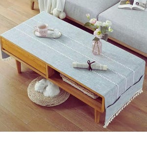 May include: A light blue rectangular table runner with white stitched lines and fringe trim. The runner is on a wooden coffee table with a drawer and a lower shelf. A small pink teacup and a vase of flowers are on the table.