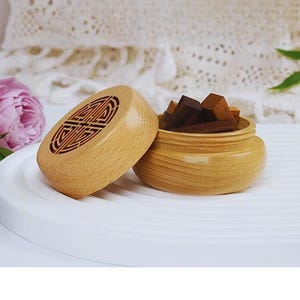 Op de afbeelding: Een houten parfumcontainer met een decoratief deksel, naast een kleine houten kom gevuld met donkerbruine houten blokken. Het deksel heeft een gesneden ontwerp. De tekst "ADD YOUR OWN PERFUME" staat bovenaan.