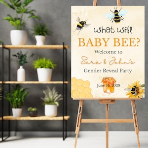May include: A wooden easel displays a sign with the text "What will BABY BEE?" and "Welcome to Sara & John's Gender Reveal Party." The sign features bee and honeycomb illustrations, with the date June 16, 2028. The background includes potted plants.