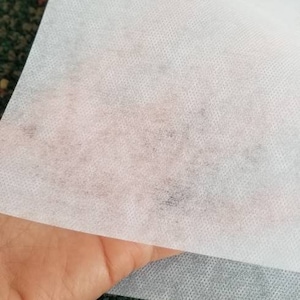 May include: A white, non-woven fabric sheet with a slightly textured surface. The sheet is folded in half and held by a hand.