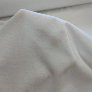 May include: White fabric with a smooth, slightly textured surface. The fabric is folded and draped, showing its soft and drapey quality.