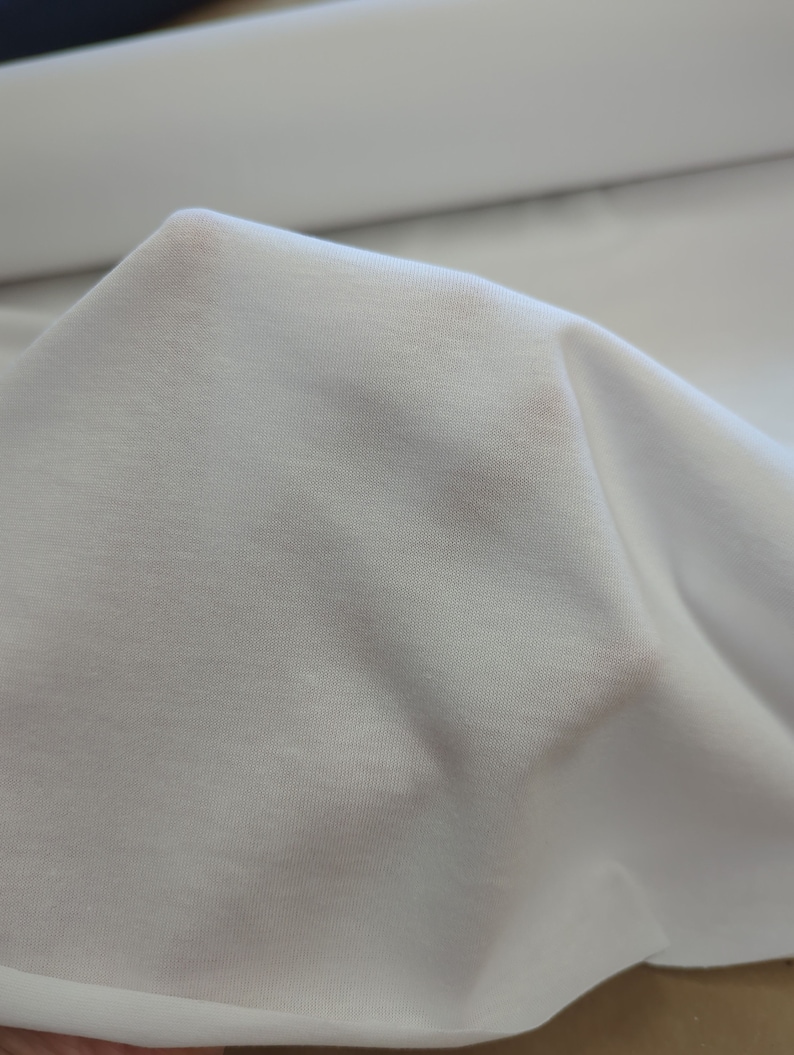 May include: White fabric with a smooth, slightly textured surface. The fabric is folded and draped, showing its soft and drapey quality.