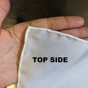 Pode incluir: Um quadrado de tecido branco com uma borda costurada. O texto "TOP SIDE" est&aacute; impresso no tecido.