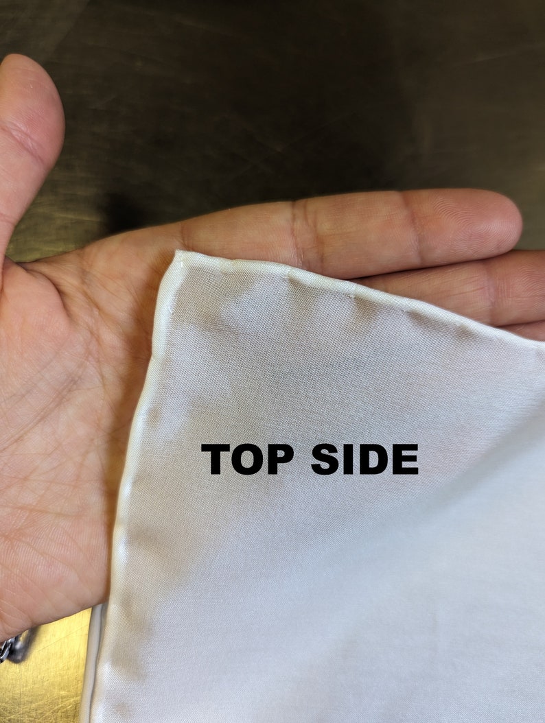 Pode incluir: Um quadrado de tecido branco com uma borda costurada. O texto "TOP SIDE" est&aacute; impresso no tecido.