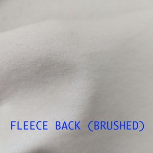 Pu&ograve; includere: Primo piano di un tessuto bianco con una texture morbida e spazzolata. Il testo "FLEECE BACK (BRUSHED)" &egrave; stampato sul tessuto.