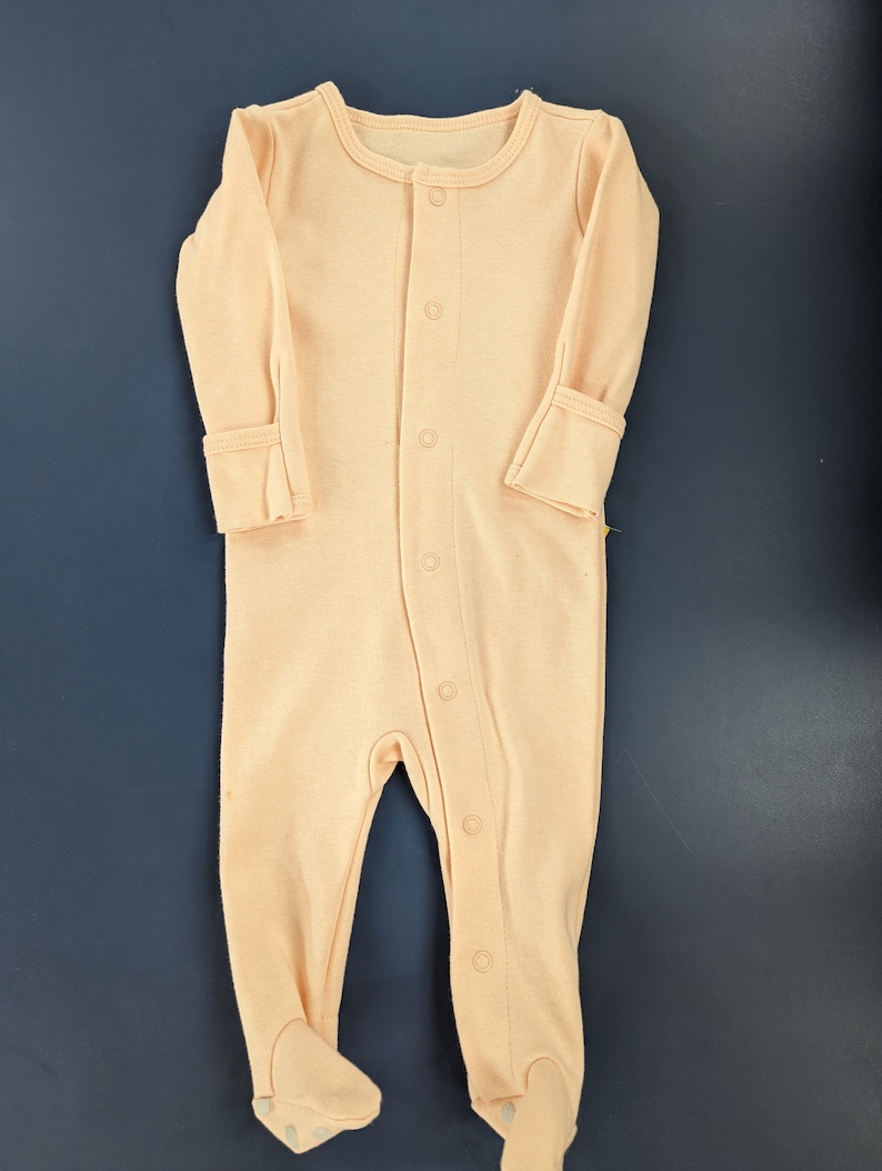 May include: A peach-colored baby onesie with long sleeves and snap buttons down the front.