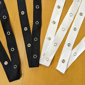 May include: Close-up of black and white fabric straps with evenly spaced metal snap closures. The straps are arranged diagonally against a light brown wooden surface. The black straps have a herringbone pattern.