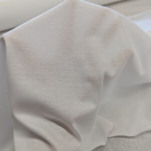 May include: White fabric with a smooth, slightly textured surface. The fabric is folded and draped, showing its soft and drapey quality.