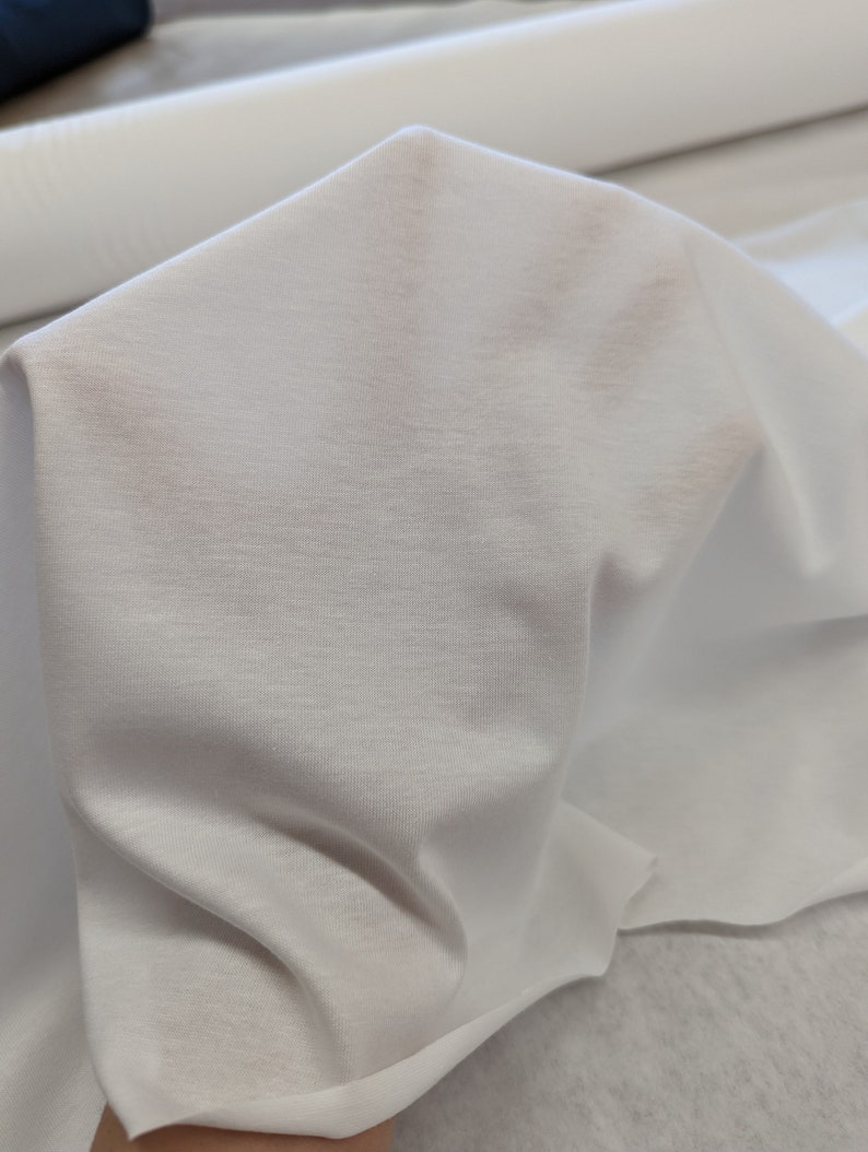 May include: White fabric with a smooth, slightly textured surface. The fabric is folded and draped, showing its soft and drapey quality.