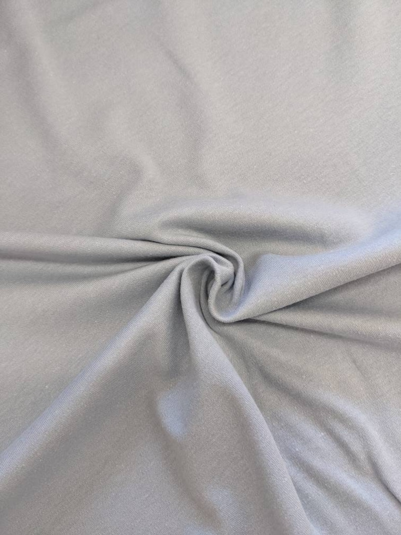 May include: A close-up of a light gray fabric with a smooth texture. The fabric is folded and draped, showing its soft and drapey quality.