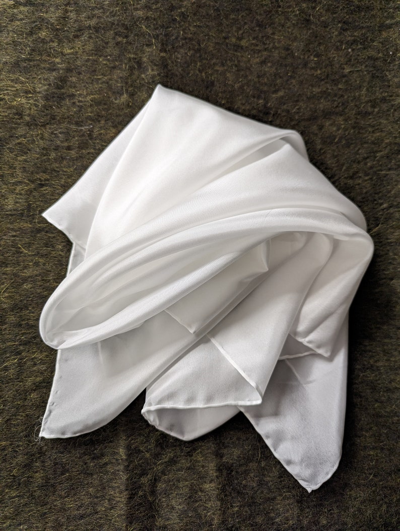 Pode incluir: Um len&ccedil;o de seda branco dobrado em forma de tri&acirc;ngulo. O len&ccedil;o &eacute; liso e tem uma textura macia e fluida.
