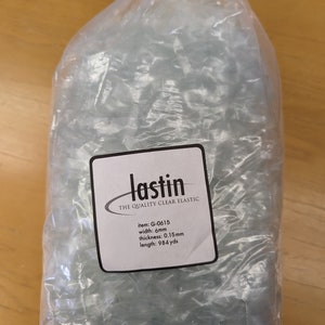 May include: A roll of clear elastic cord packaged in a transparent plastic bag. A white label on the front reads: lastin THE QUALITY CLEAR ELASTIC item: G-0615 width: 6mm thickness: 0.15mm length: 984 yds. The elastic is coiled tightly inside the bag.