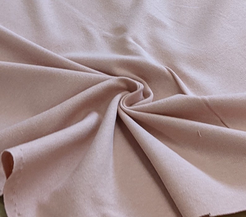 May include: A close-up of a soft, pale pink fabric with a smooth texture. The fabric is folded and draped, showing its natural drape and flow.