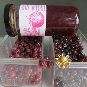 May include: Two clear plastic containers filled with red and white glass beads. A glass jar of raspberry jam with a pink and white label is laying on top of the containers. The label reads "Daybreak Kitchen Raspberry After-Pink".
