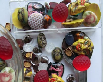 Puede incluir: Una colección de botones de vidrio y plástico coloridos, que incluyen una variedad de formas, tamaños y diseños. Algunos botones presentan un patrón floral, mientras que otros tienen un diseño geométrico. También hay varios botones con un diseño de cara. Los botones están dispuestos en un recipiente de plástico transparente y sobre una superficie blanca.