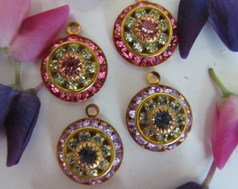 May include: Four gold-tone pendants with colorful crystal embellishments. Each pendant features a round, multi-colored crystal surrounded by smaller crystals. The pendants are arranged on a white surface with pink and purple flowers.