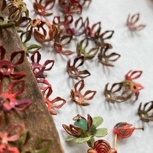 Puede incluir: Primer plano de varias tapas de cuentas metálicas con forma de flor en tonos rojos, verdes y morados. Algunas están agrupadas sobre una superficie de madera, mientras que otras están dispersas sobre un fondo blanco. Se incluye una moneda como referencia de tamaño.