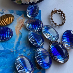 May include: A small bowl filled with blue glass imitation oval stones. The bowl is decorated with floral designs and gold accents. The stones have a striated pattern and a clear, iridescent finish. The label on the bag reads "Glass Imitation Stones", "No. 6692", "Ovals", "Size 18/13", "1/2 gross", and "col. 80".