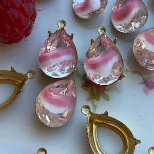 May include: Assortment of pink and white glass teardrop-shaped embellishments with gold metal settings. The embellishments have a swirl pattern and are arranged on a white surface with a floral design.