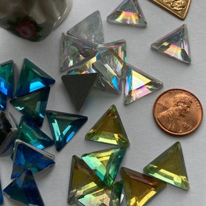 May include: Assortment of iridescent triangle-shaped glass rhinestones in various colors, including blue, green, and gold. The rhinestones are scattered on a white surface, with a penny for scale.