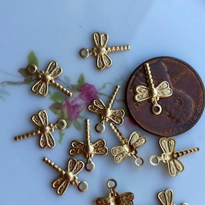 May include: A collection of ten gold-toned dragonfly charms with a beaded tail. The charms are arranged on a white plate with a floral pattern.