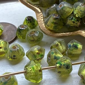 May include: A collection of faceted, translucent lime green glass beads with dark green and black internal flecks. Some beads are loose, while others are strung on a thin gold-toned wire. A copper-colored coin is visible near the beads.