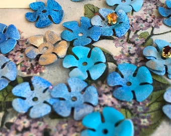 May include: A collection of blue fabric flower embellishments with a five-petal design. The flowers have a textured, slightly distressed look and are scattered on a floral background.