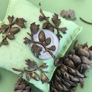 May include: A green fabric sachet with a drawstring closure decorated with metallic pine cone and branch embellishments. The sachet is surrounded by pine cones on a green background.