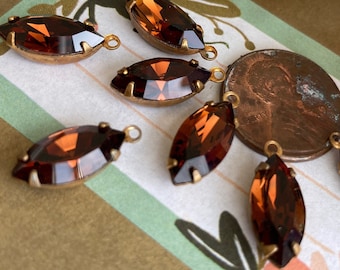 May include: A collection of eight brown marquise-shaped glass rhinestones with gold-toned metal settings. The rhinestones have a faceted design and are arranged on a patterned surface.