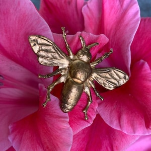 May include: A gold-colored metal bee figurine on a pink flower. The bee has detailed wings and legs. The flower is a gladiolus.