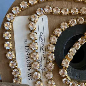 Puede incluir: Un carrete de cadena dorada con cristales de imitación transparentes. La etiqueta dice "SWAROVSKI TM" y "Item # 2088  Crystals  Foiled  Clear".