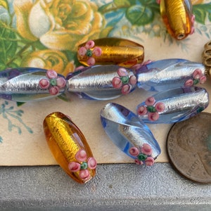 Op de afbeelding: Assortiment van ovale glazen kralen met zilveren en gouden folie accenten. De kralen zijn versierd met roze en groene bloemen. Een kleine kom gevuld met vergelijkbare kralen staat op de achtergrond.