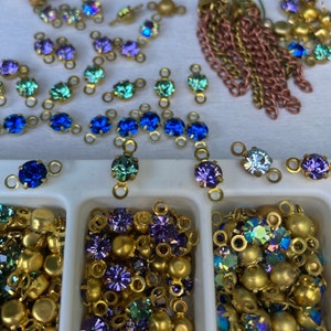 May include: Assortment of gold-toned metal connector charms with various coloured glass rhinestones. The charms are arranged in rows on a white surface and in a small white tray with compartments. The rhinestones are in shades of blue, green, purple, and iridescent.