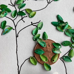 May include: Green metal leaf-shaped charms with a hole in the center, scattered around a US penny coin. The coin is surrounded by a larger group of charms.