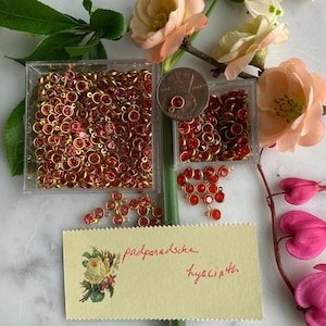 May include: Two small containers filled with red glass beads with gold accents. The containers are on a white marble surface with a handwritten note that says "padparadscha hyacinth".