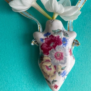 Pode incluir: Um vaso de cerâmica branco com um padrão floral, com flores rosa, amarelas e azuis. O vaso está pendurado em uma fita dourada e azul e tem flores brancas dentro.